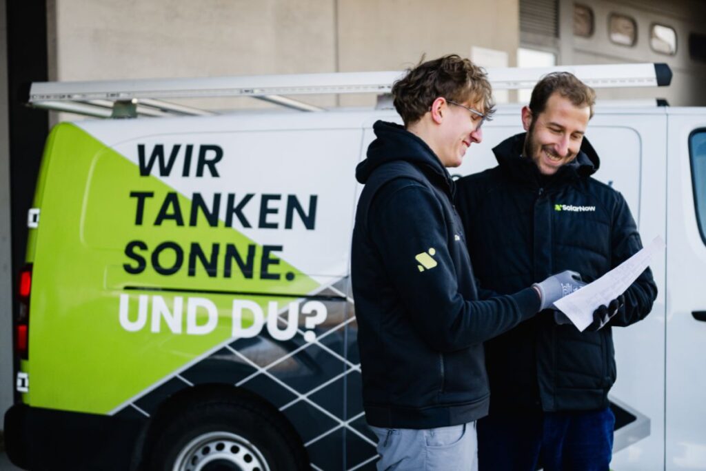 Zwei SolarNow-Mitarbeiter in Arbeitskleidung besprechen Unterlagen vor einem Firmenfahrzeug mit der Aufschrift „Wir tanken Sonne. Und du?“.