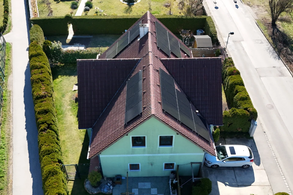 PV Anlage Schrägdach Maria Enzersdorf SolarNow auf einem hellgrünen Einfamilienhaus mit braunem Ziegeldach, weißem PKW in der Garageneinfahrt und hohen Hecken, installiert von SolarNow