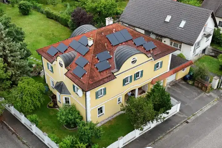 PV Anlage auf rotem Schraegdach Einfamilienhaus Baden bei Wien zeigt eine Luftaufnahme einer gelben Villa mit verwinkeltem Walmdach und zahlreichen schwarzen Solarmodulen inmitten eines grünen Gartens, installiert von SolarNow