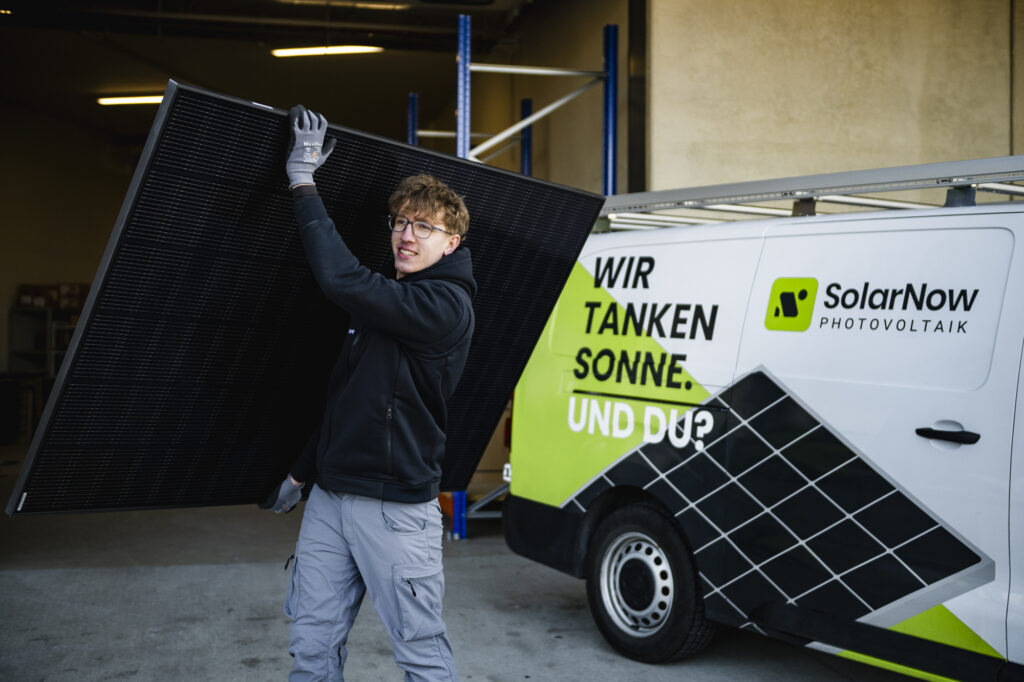 Mitarbeiter von SolarNow trägt Solarpanel vor Firmenfahrzeug mit Slogan ‚Wir tanken Sonne. Und du?‘ – Photovoltaik-Montage in Österreich