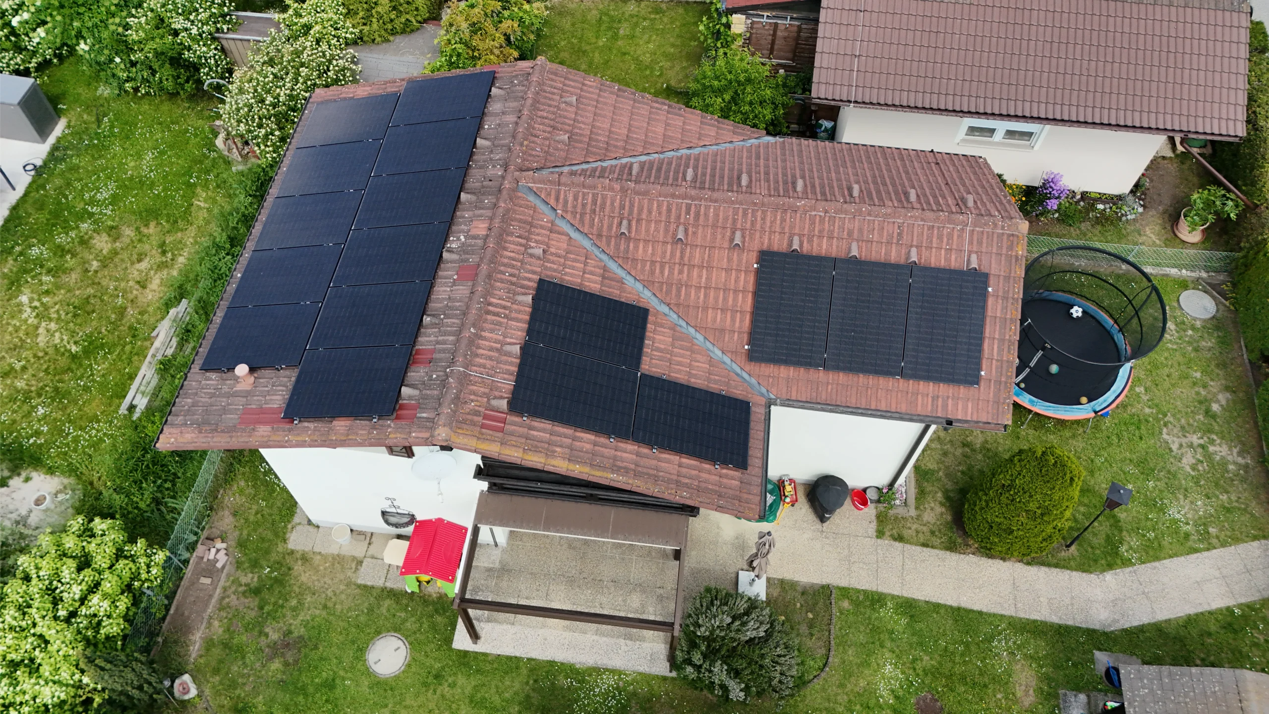 PV-Anlage Kleingarten Favoriten zeigt eine Luftaufnahme eines modernen Kleingartenhauses mit anthrazitfarbenem Satteldach und installierter Photovoltaikanlage, angrenzend an eine Terrasse und einen Garten mit Pool, präsentiert von SolarNow